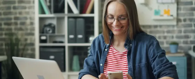 Competitive Compensation: a woman sitting at a table using smartphone at work swiping looking at screen and smiling