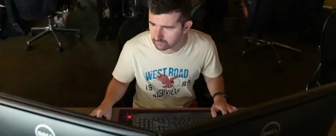 DLT Security: Man in casual t-shirt working from an office on dual monitors.