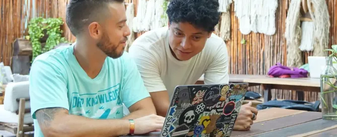 glossary: man in teal crew neck t-shirt sitting beside man in white crew neck t-shirt Two digital nomads working and collaborating remotely on a computer outside.