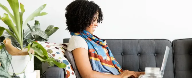 Trabalhar Remoto para Empresas Americanas / Performance Improvement Plan: A woman smiles as she relaxes on a stylish gray sofa, working on her laptop. Potted plants decorate the bright room, and a book and a cup sit on a coffee table. A relaxing remote workspace.