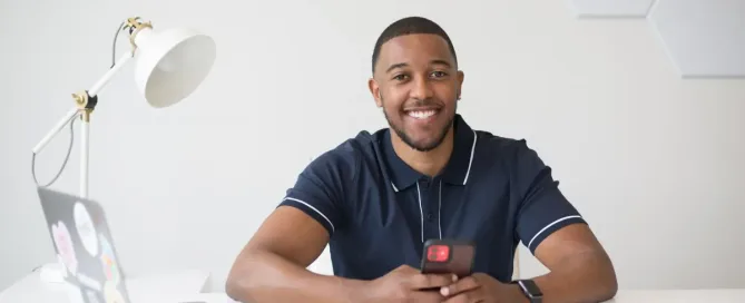 hire swift developers: young black software engineer smiles at camera, iphone in hand and imac at his side