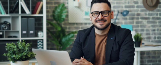 a man sitting at a table with a laptop, looking at camera smiling