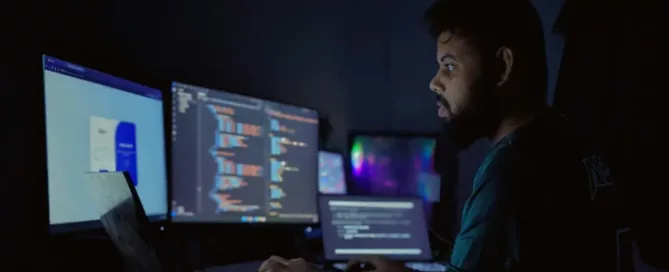 time-to-productivity: A software developer working late at night in a dimly lit room, surrounded by multiple computer screens displaying lines of code. The scene captures the focused and immersive environment of a tech professional engaged in programming and software development, showcasing a modern multi-screen setup ideal for coding and development tasks.