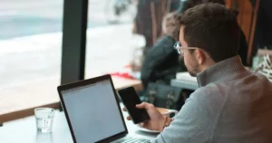 Mistakes When Hiring Software Developers; man sitting near table with laptop and smartphone near window